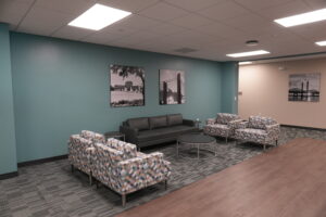 Lounge area with teal walls, a dark gray sofa, and patterned armchairs arranged around round tables; black-and-white wall art above the sofa and a beige wall with a bridge photo in the background