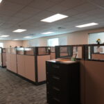 Open-plan office with beige cubicles, filing cabinets, and large windows; an illuminated EXIT sign is visible on the left.