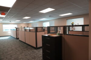 Open-plan office with beige cubicles, filing cabinets, and large windows; an illuminated EXIT sign is visible on the left.