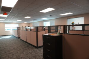 Open-plan office with row of beige cubicles, filing cabinet, and an illuminated exit sign in the ceiling.