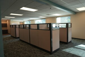 Open-plan office with beige cubicles, frosted glass panels, and bright ceiling lights along a grid ceiling.