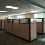 Open-plan office with beige cubicles and partitioned workstations under bright ceiling lights.