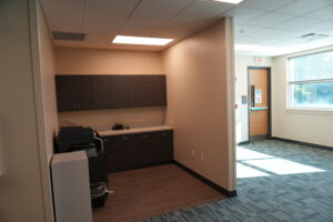 Office kitchenette with dark brown cabinets, a counter, and a printer in the foreground; exit door and window to the right.
