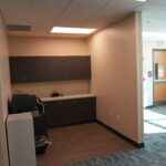 Office kitchenette with dark brown cabinets along a beige wall, a printer on the counter, and an exit door visible down a hallway.