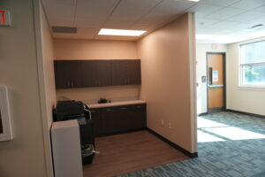 Office kitchenette with dark brown cabinets along a beige wall, a printer on the counter, and an exit door visible down a hallway.