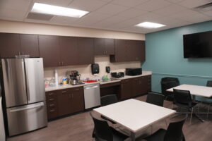 Office break room with dark brown cabinets, stainless refrigerator, coffee station, and a wall-mounted TV on a teal wall.