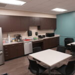 Break room/kitchen area with brown cabinets, stainless refrigerator, coffee station, microwave, and teal accent wall with a mounted TV on the right.