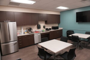 Break room/kitchen area with brown cabinets, stainless refrigerator, coffee station, microwave, and teal accent wall with a mounted TV on the right.