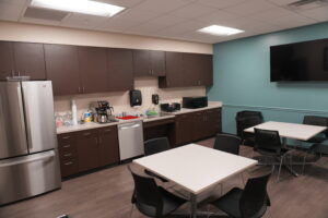 Office break room with brown cabinets, stainless refrigerator, coffee station, and a mounted flat-screen TV on a teal wall.