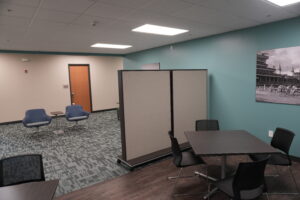 Office meeting area with teal accent wall, gray room dividers, and blue lounge chairs around small tables under a grid ceiling.