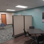 Office lounge with teal wall, divider panels, and cluster of blue swivel chairs around small tables.