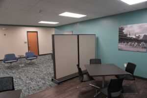 Office lounge with teal wall, divider panels, and cluster of blue swivel chairs around small tables.