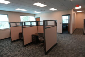 Open-plan office with beige cubicles, desks, and mesh chairs under bright ceiling lights, plus an exit sign in a hallway to the right.