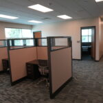 Open-plan office with beige cubicles, gray desks, and a patterned carpet under bright ceiling lights.