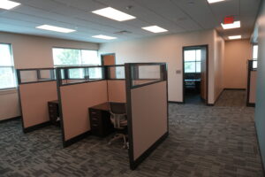 Open-plan office with beige cubicles, gray desks, and a patterned carpet under bright ceiling lights.