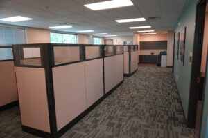 Open-plan office with a row of beige cubicles and frosted glass panels along a carpeted walkway under bright ceiling lights.