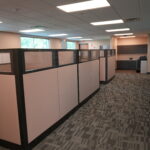 Open-plan office with row of beige and gray cubicles, frosted glass panels, and a kitchenette in the back under fluorescent lights.