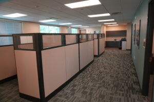 Open-plan office with row of beige and gray cubicles, frosted glass panels, and a kitchenette in the back under fluorescent lights.