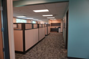 Office hallway with row of beige cubicles, glass panels, and overhead fluorescent lights
