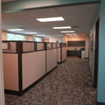 Office hallway with cubicles on left and teal wall on right, overhead fluorescent lights, and a kitchenette in the distance