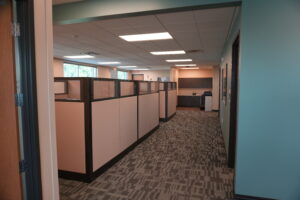 Office hallway with cubicles on left and teal wall on right, overhead fluorescent lights, and a kitchenette in the distance