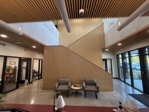 Modern lobby interior with a diagonal wood panel partition, two gray armchairs, and a small round table under a wooden slat ceiling.