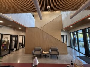 Lobby area with wooden slat ceiling, glass railings, and a central wooden divider; two gray chairs and a small round table in front of the divider.
