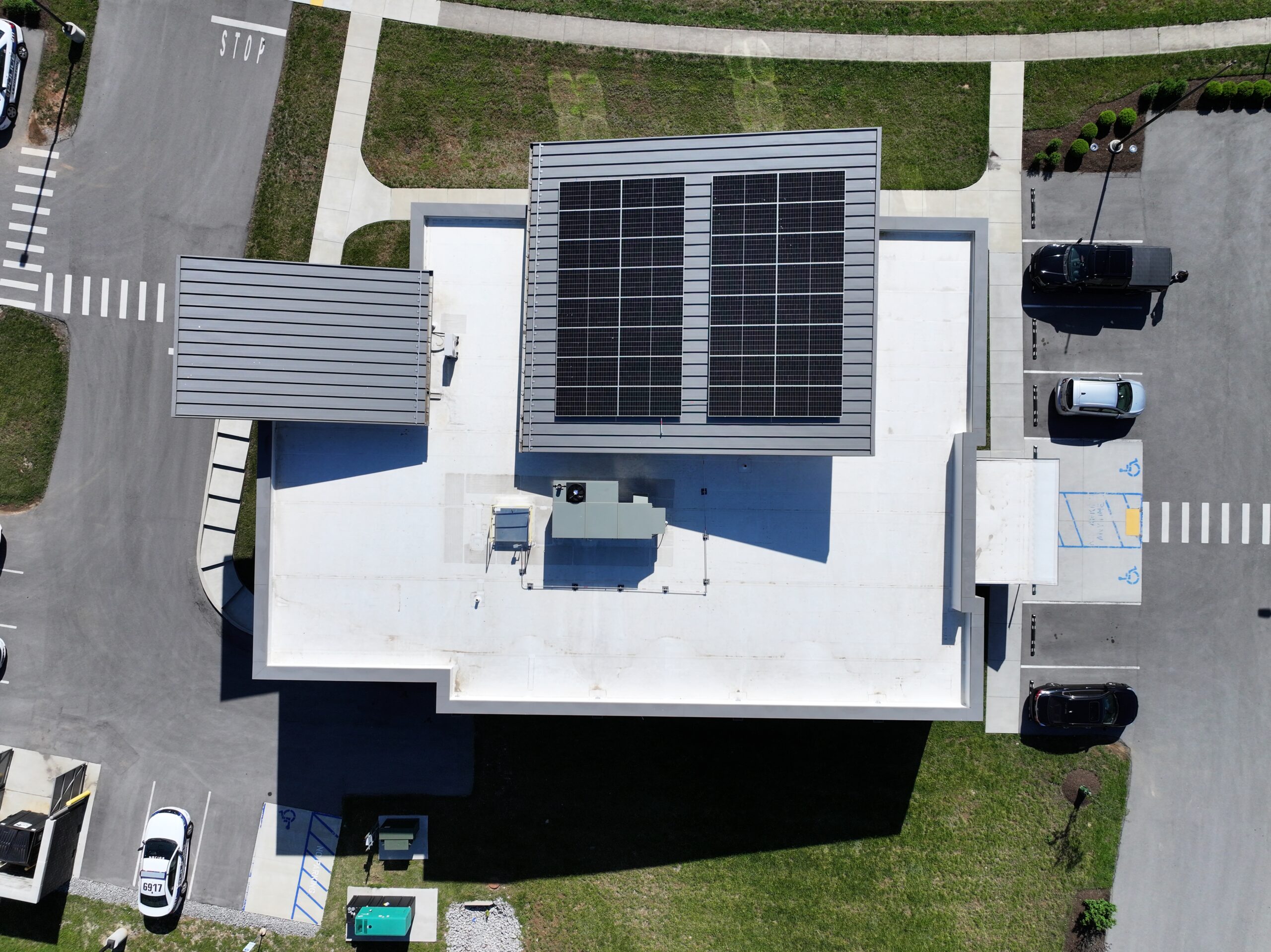 Aerial view of a building with solar panels on the roof, surrounded by a concrete lot, walkways, and green lawn areas.