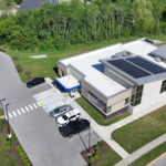 Aerial view of a modern, flat-roofed building with solar panels, parking lot, and surrounding green space.