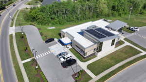 Aerial view of a modern, flat-roofed building with solar panels, parking lot, and surrounding green space.