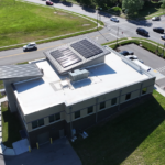 Aerial view of a single-story commercial building with two solar-panel arrays on the flat roof, surrounded by roads and green space with cars nearby in a parking lot.