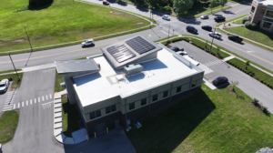 Aerial view of a single-story commercial building with two solar-panel arrays on the flat roof, surrounded by roads and green space with cars nearby in a parking lot.