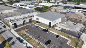 Aerial view of a modern white and gray commercial building with a large paved parking lot and several parked cars, surrounded by industrial buildings and a railroad track in the background.