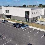 Aerial view of a modern two-story office building with a large parking lot and several parked cars, including a designated handicap space near the entrance.