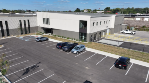 Aerial view of a modern two-story office building with a large parking lot and several parked cars, including a designated handicap space near the entrance.