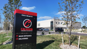 Calhoun Construction Services sign with logo and arrows for Main Entrance and Deliveries, in front of a modern office building on a sunny day